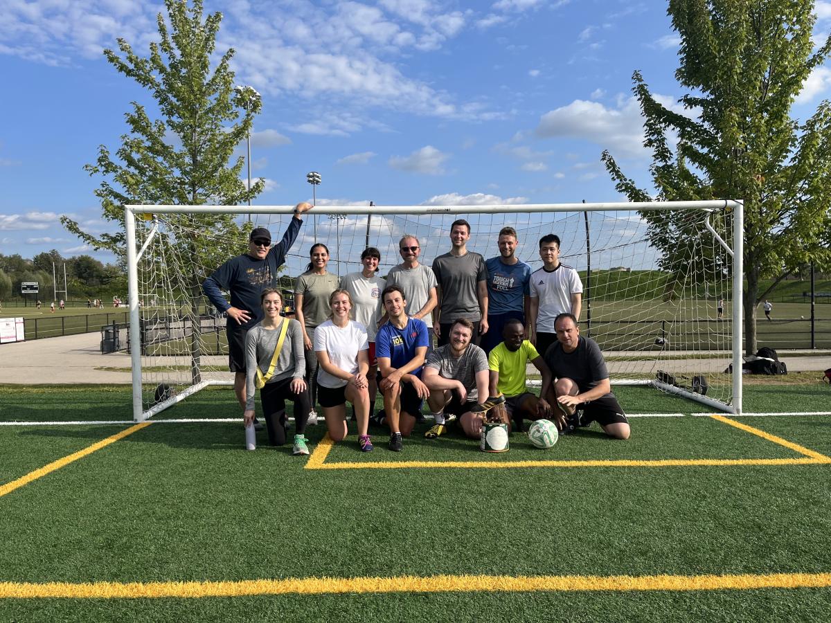 Picture showing 13 people wearing sports clothes and standing inside a soccer goal, all looking at the camera and smiling. 