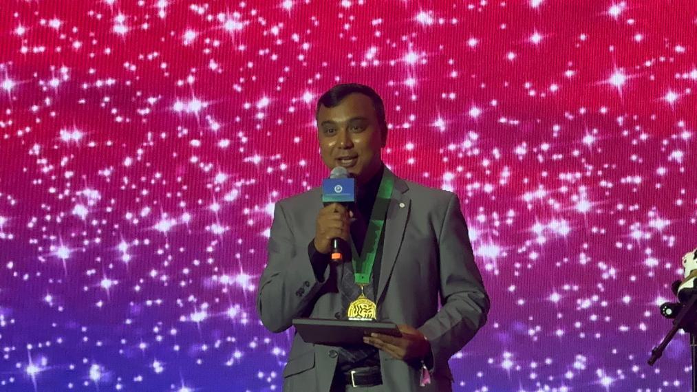 Dr. Asim Biswas smiling and talking into a microphone with a medal around his neck, standing in front of a very sparkly background. 