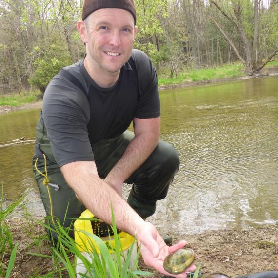 Picture of Dr. Ryan Prosser kneeling at the side of a river holding a mollusc shell towards the camera
