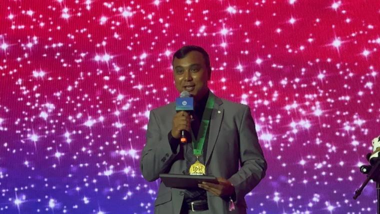 Dr. Asim Biswas smiling and talking into a microphone with a medal around his neck, standing in front of a very sparkly background. 