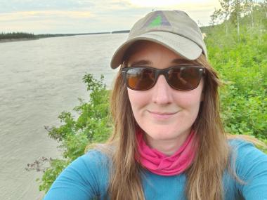 Portrait of Dr. Catherine Dieleman. She is wearing a hat, sunglasses, a pink scarf and a blue shirt and is standing next to a waterway