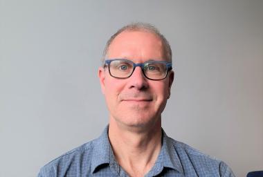 Headshot of Simon Lachance wearing blue glasses, a blue shirt and sitting in front of a plain grey background.